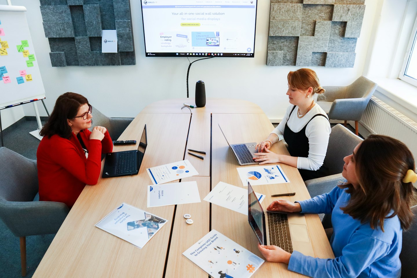 Three women collaborate around a table with laptops in an office.